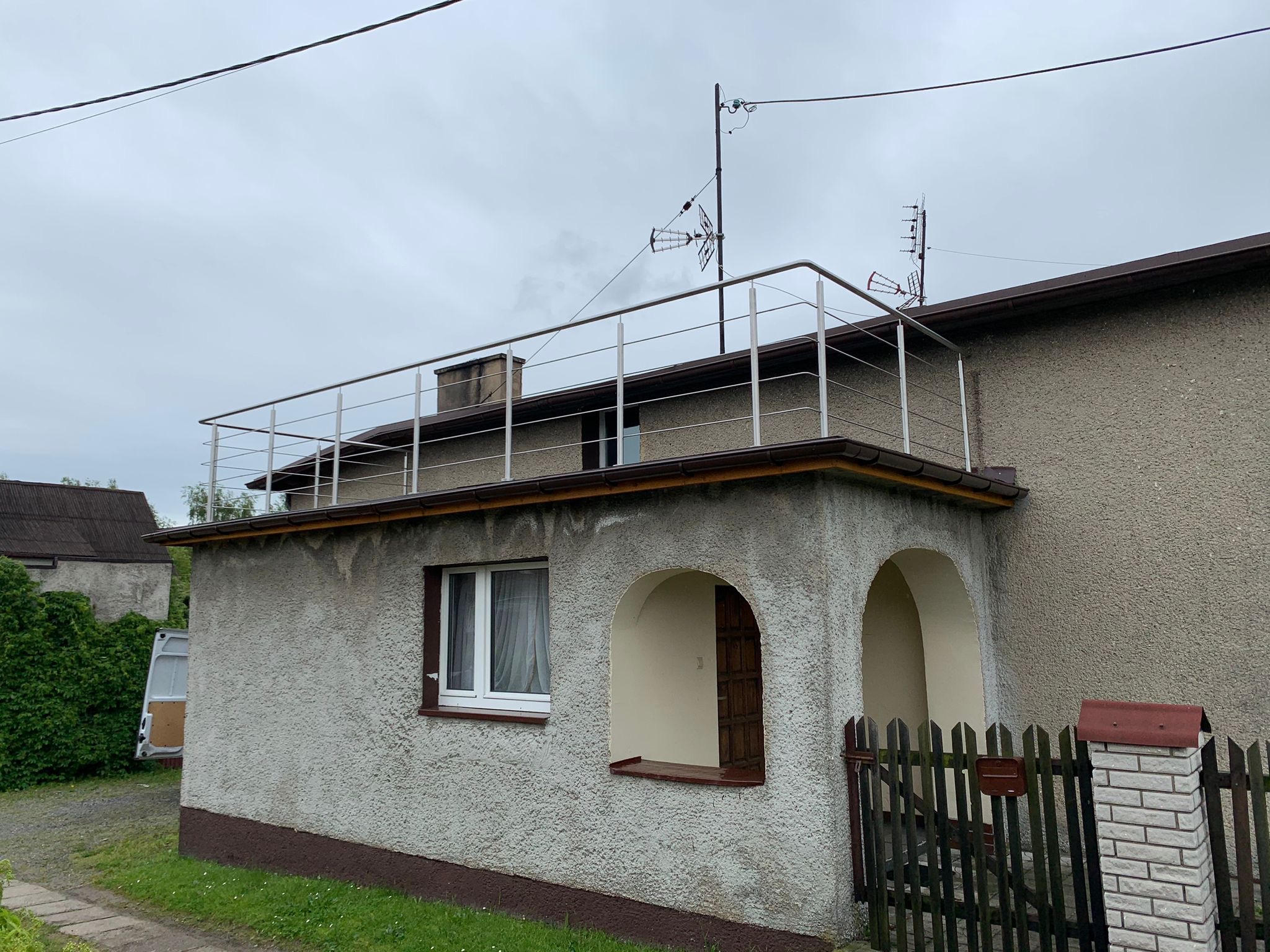 Balkon z balustradą ze stali nierdzewnej na parterowym budynku z charakterystyczną elewacją typu baranek, widoczne anteny na dachu, pochmurne niebo w tle.