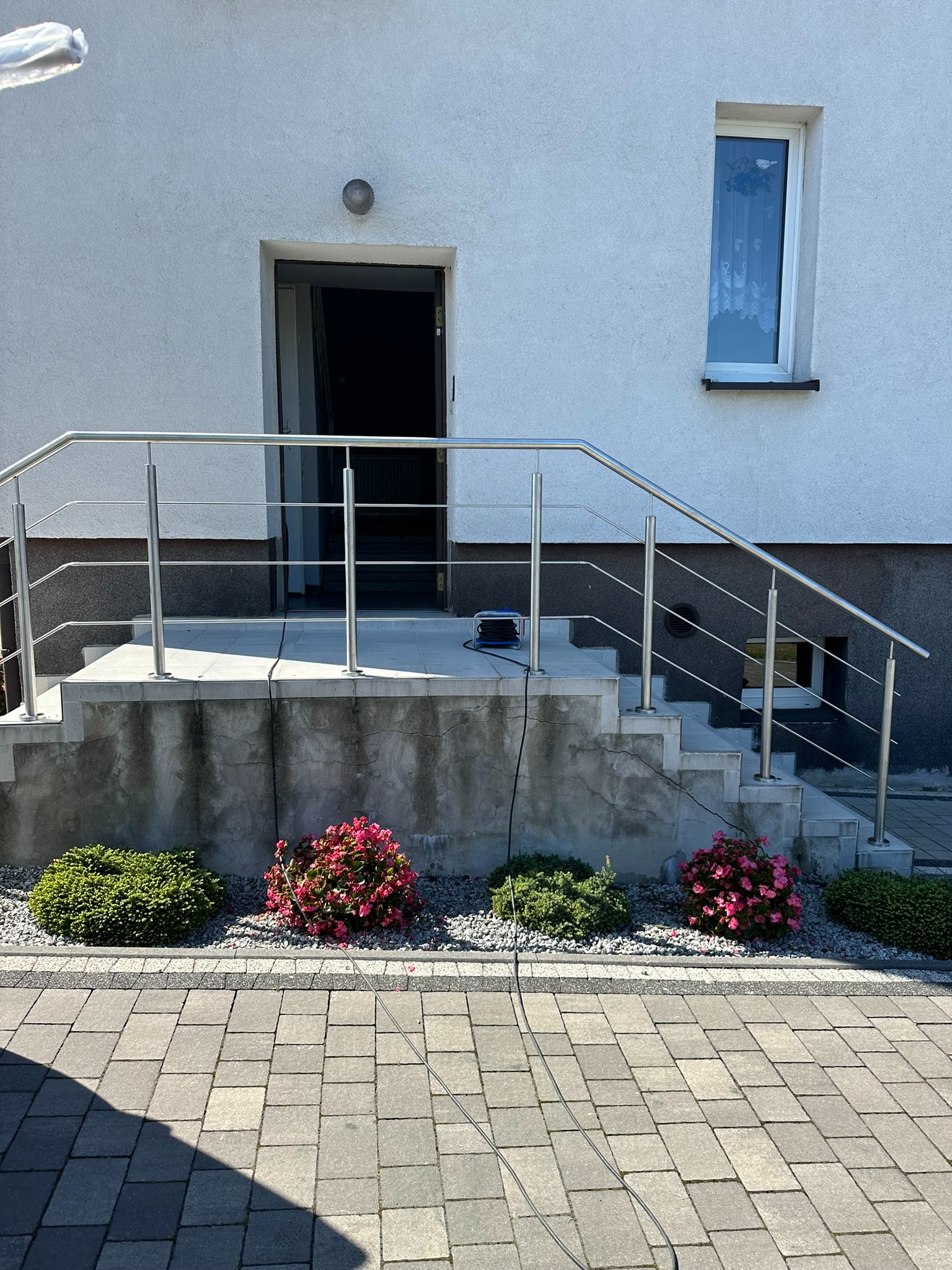 Zewnętrzne schody wejściowe do domu z balustradą ze stali nierdzewnej, widoczne kwiaty i kostka brukowa, kabel od urządzenia leżący na schodach.