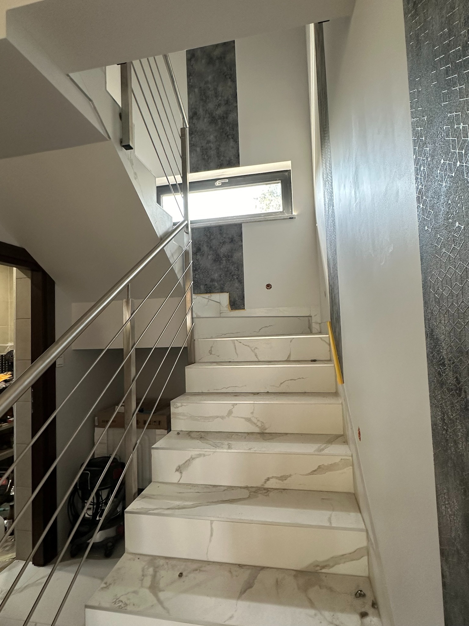 Wnętrze domu w trakcie wykańczania: Schody wyłożone białym marmurem z szarymi żyłkami, zabezpieczone metalową, nowoczesną poręczą z cienkich prętów. Widoczne okno na półpiętrze i fragment...