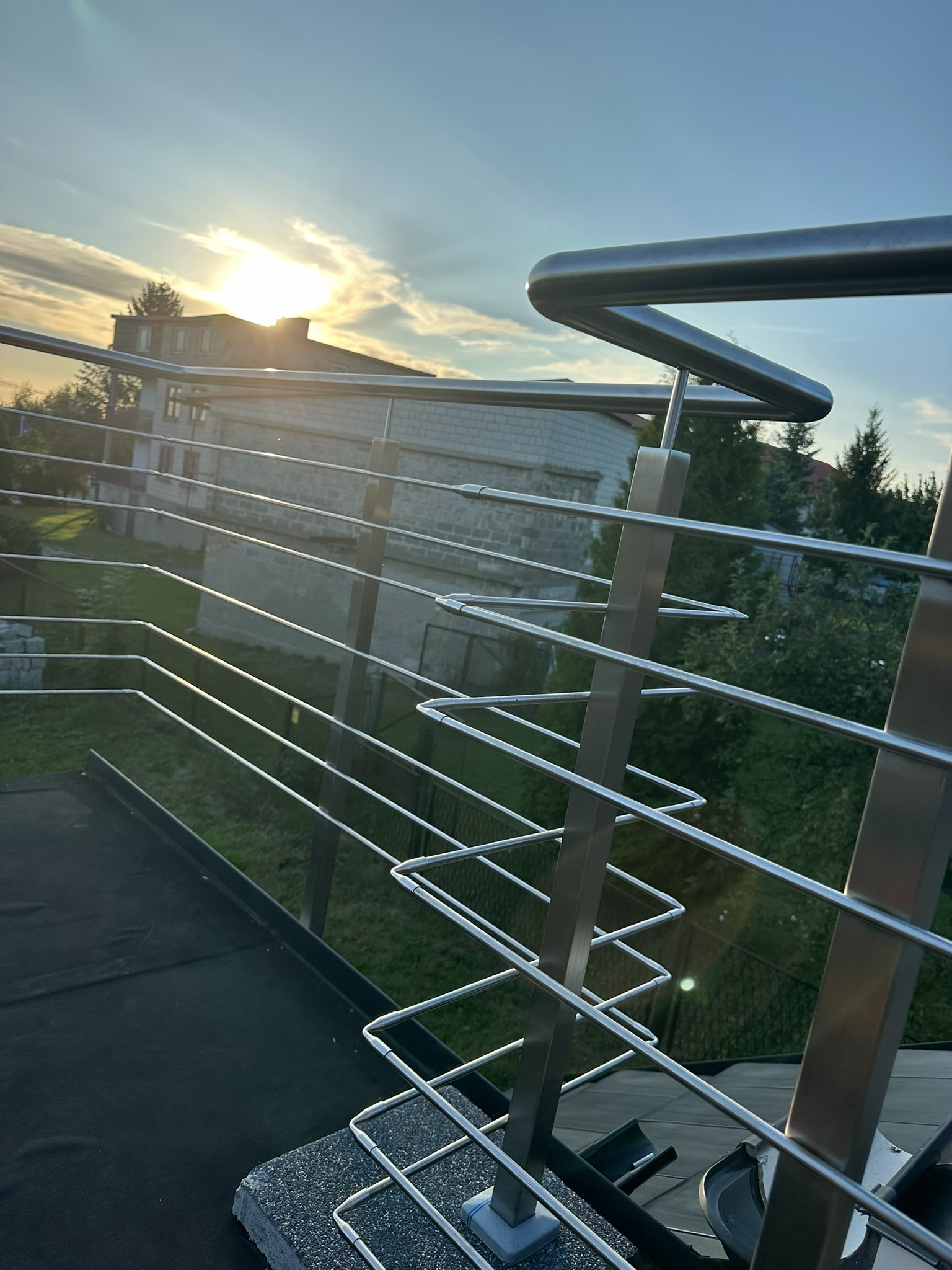 Nowoczesna balustrada ze stali nierdzewnej z poziomymi prętami, widok z góry, w tle dom i zachodzące słońce.