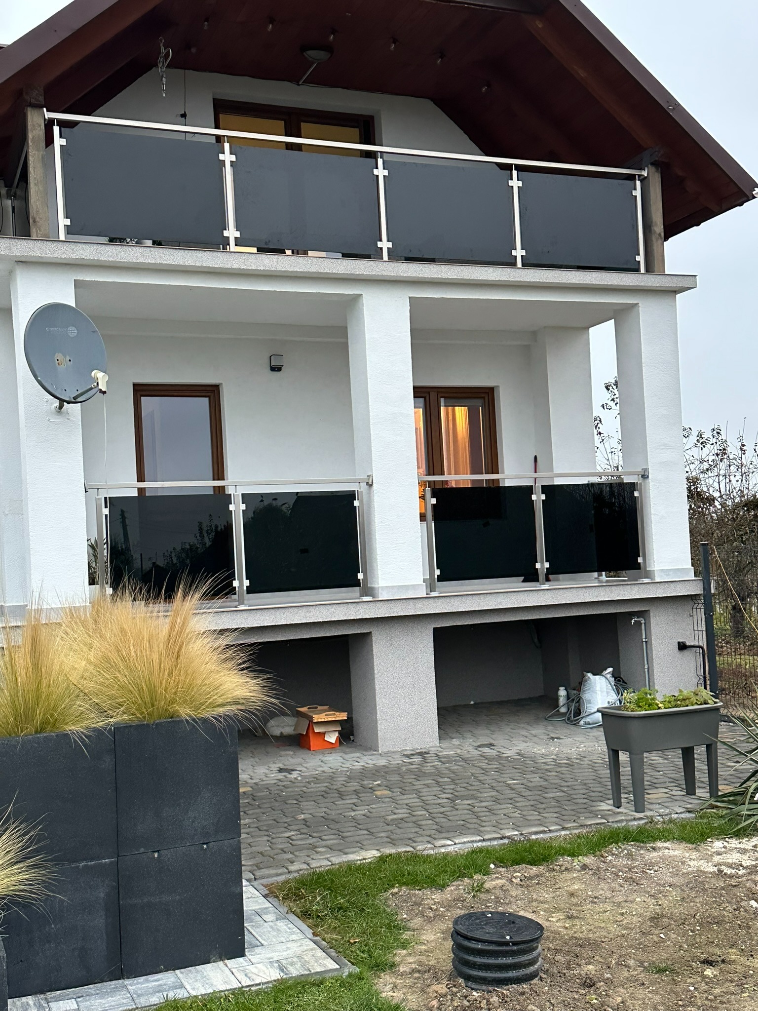 Dwupiętrowy dom z balkonami zabezpieczonymi panelami z czarnego szkła i nierdzewnymi słupkami, widok z poziomu ogrodu z dekoracyjnymi trawami w donicach.