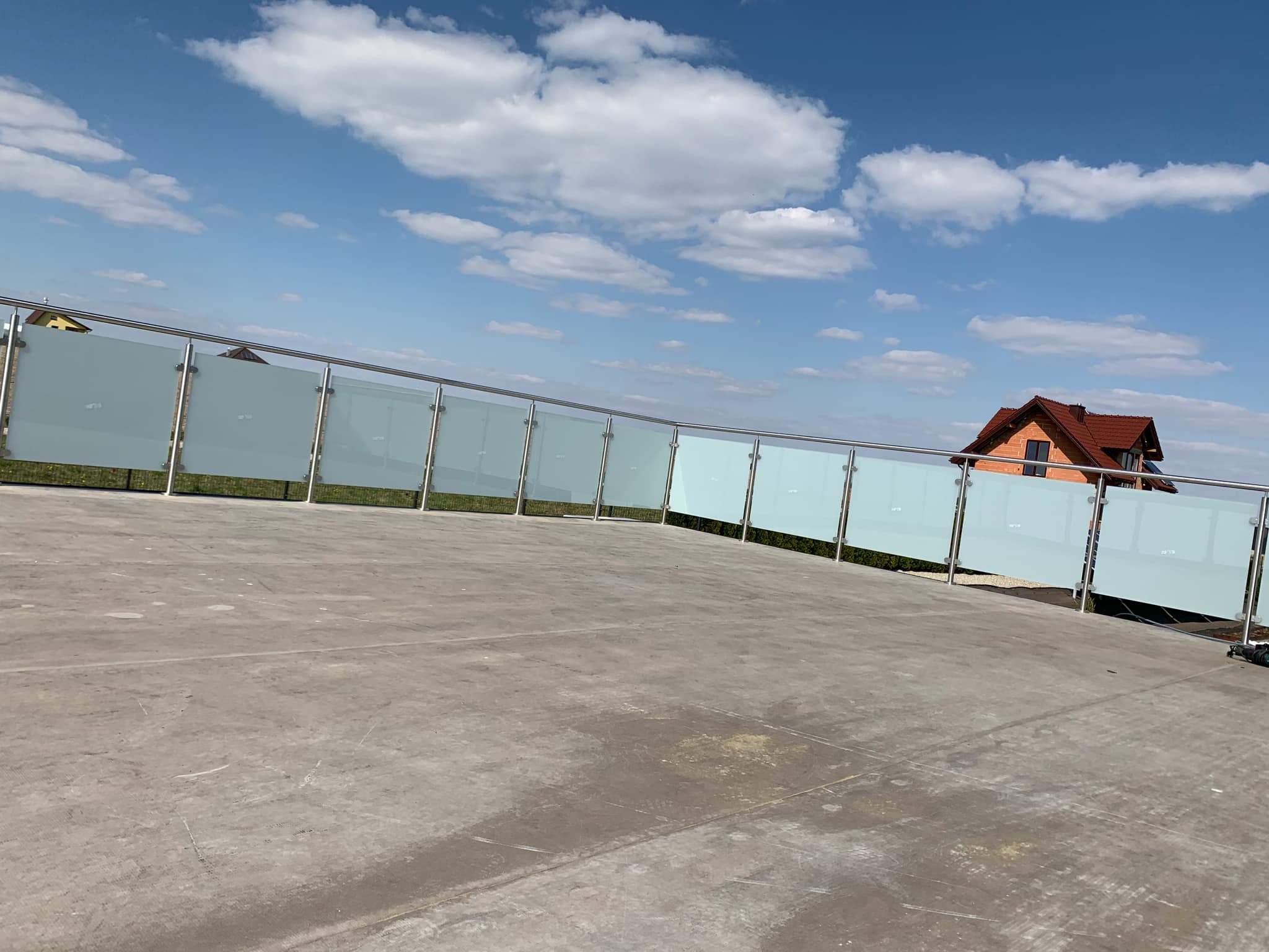 Szklana balustrada z nierdzewnymi słupkami, zamontowana na tarasie w Katowicach, widok z dołu na niebieskie niebo z chmurami i dom w tle.