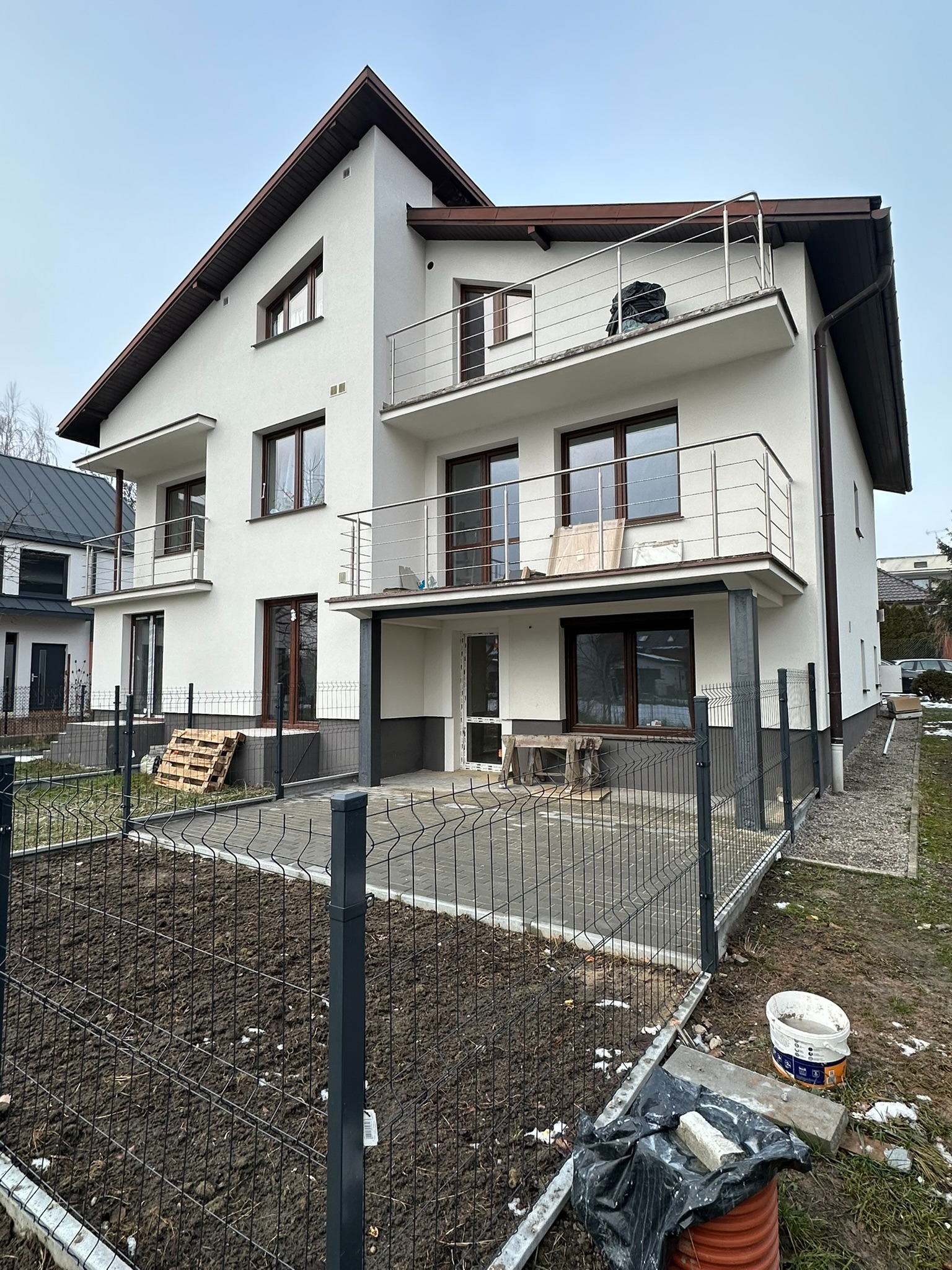 Nowoczesny dom z balkonami zabezpieczonymi metalowymi, minimalistycznymi barierkami, widok z poziomu gruntu na niezagospodarowany ogród z siatką ogrodzeniową.