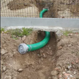 Wykonany wykop z ułożoną zieloną rurą karbowaną z metalowym złączem, widoczny mini-koparka i studzienka kanalizacyjna w tle, ziemia z gruzem.