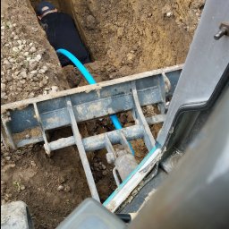 Krzyśkop - Wykop z ułożoną niebieską rurą, widok z kabiny maszyny budowlanej na pracownika w dole wykopu.
