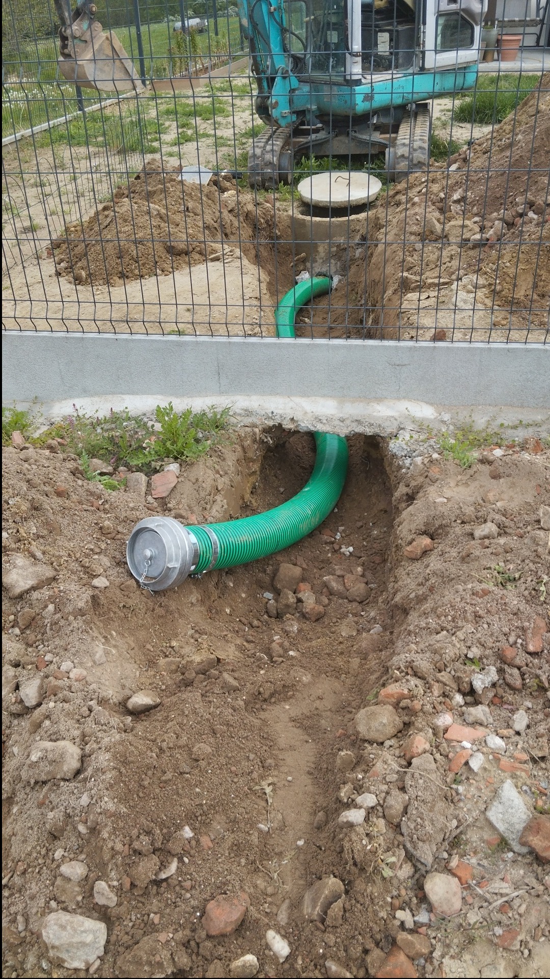 Wykonany wykop z ułożoną zieloną rurą karbowaną z metalowym złączem, widoczny mini-koparka i studzienka kanalizacyjna w tle, ziemia z gruzem.