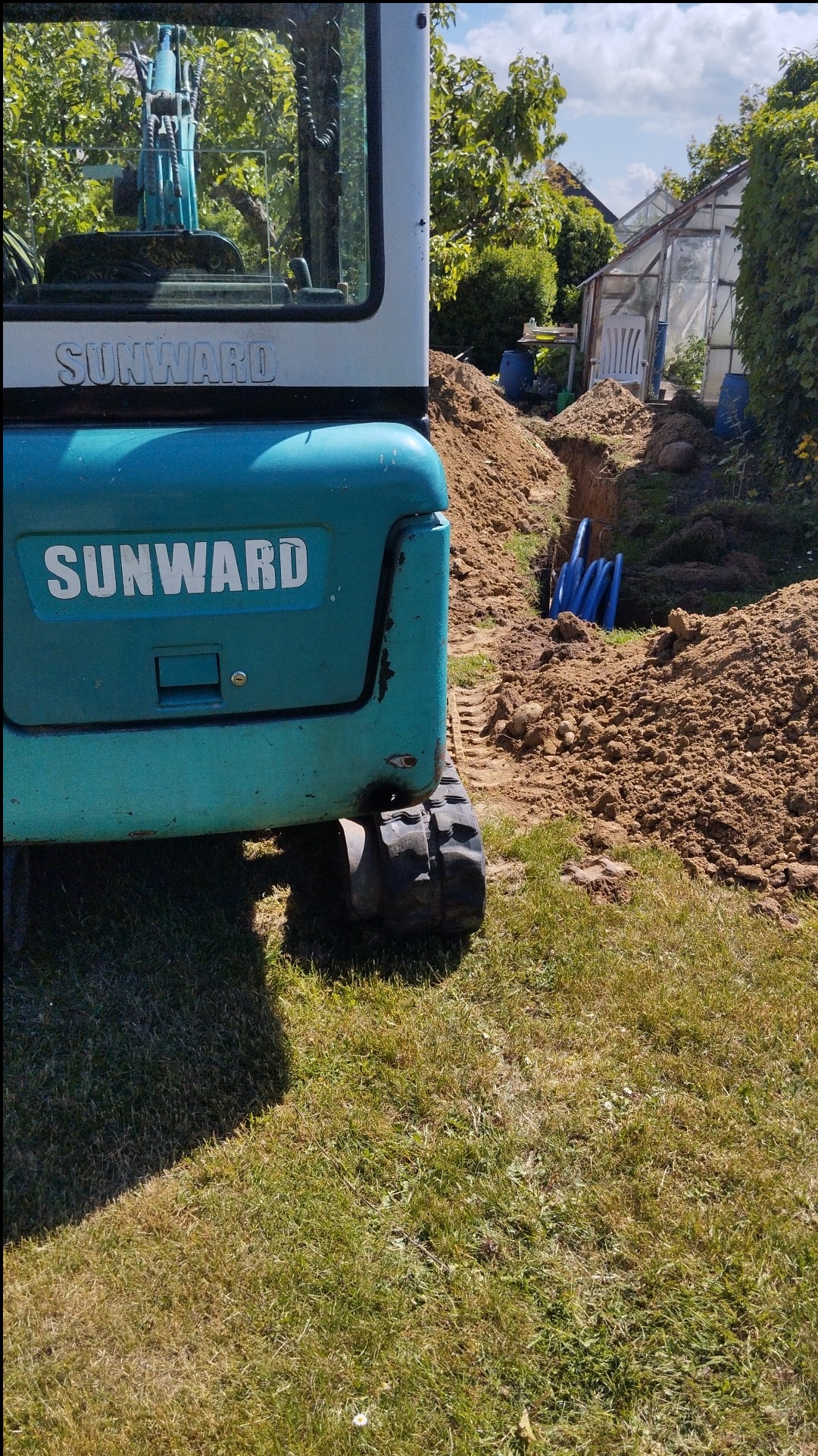 Turkusowa minikoparka marki Sunward częściowo widoczna obok wykopu z niebieskimi rurami, w tle szklarnia i zieleń ogrodu.
