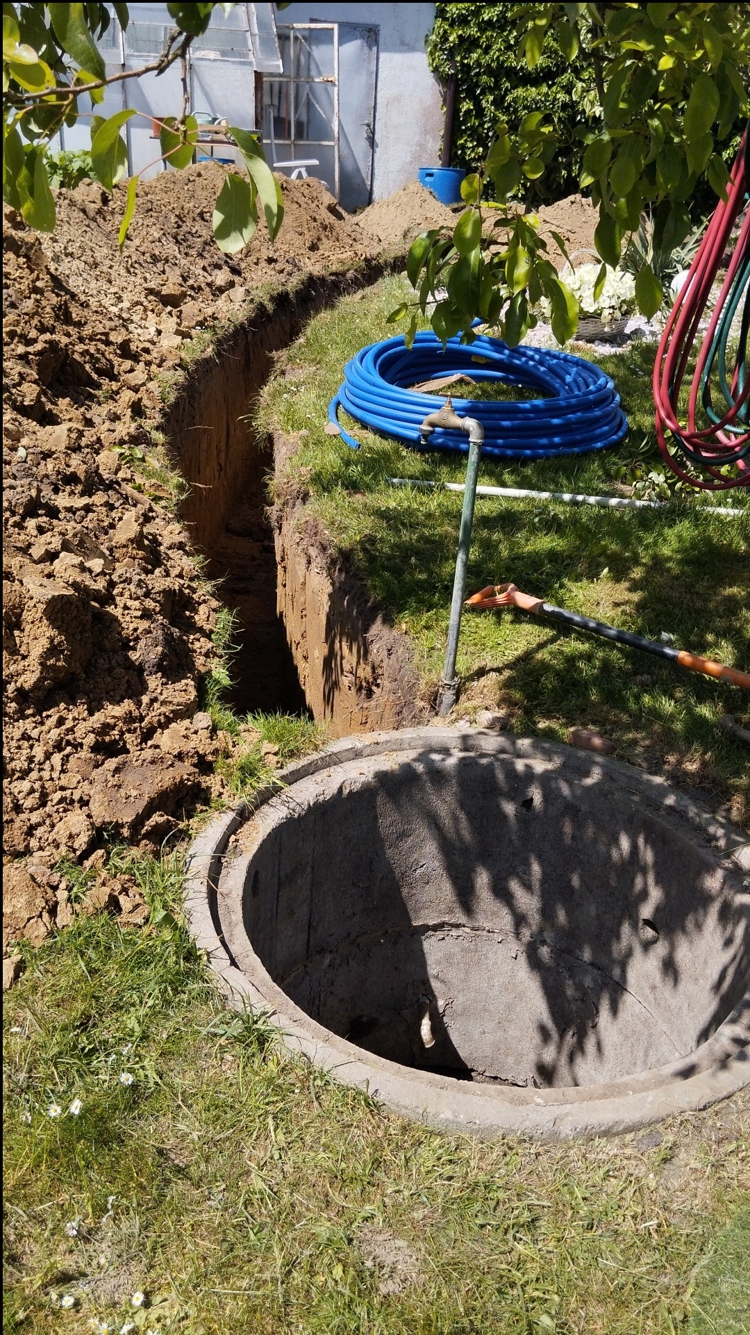 Otwarty, betonowy krąg studzienny w trawie, obok wykopany rów z widoczną stertą ziemi, niebieski zwój rur i częściowo widoczne czerwone węże w tle.