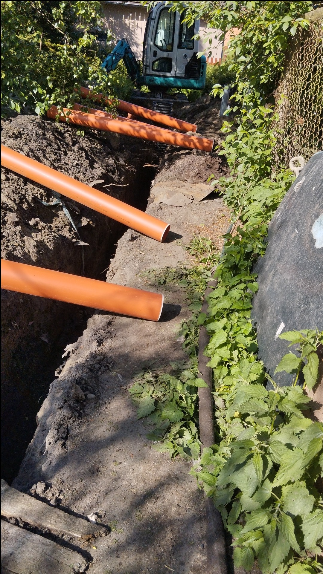 Wykopany rów z ułożonymi pomarańczowymi rurami kanalizacyjnymi, w tle widoczna mała koparka, teren częściowo porośnięty zielenią.