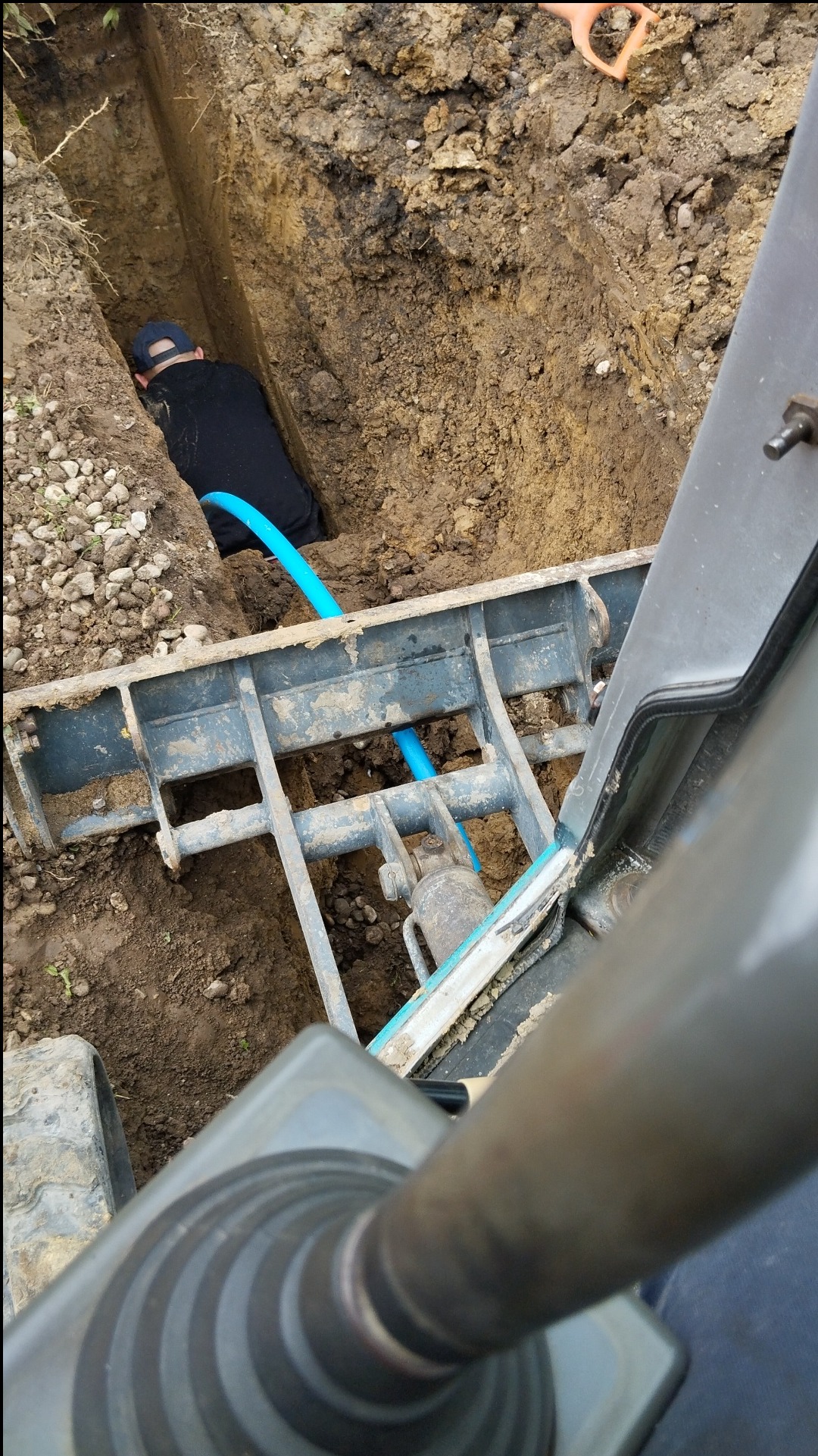 Wykop z ułożoną niebieską rurą, widok z kabiny maszyny budowlanej na pracownika w dole wykopu.
