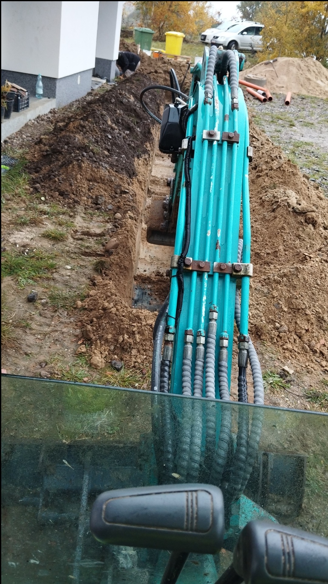 Widok z kabiny koparki na wykopany wąski rów w ziemi, praca w toku, w tle fundament domu i osoba przy wykopie.