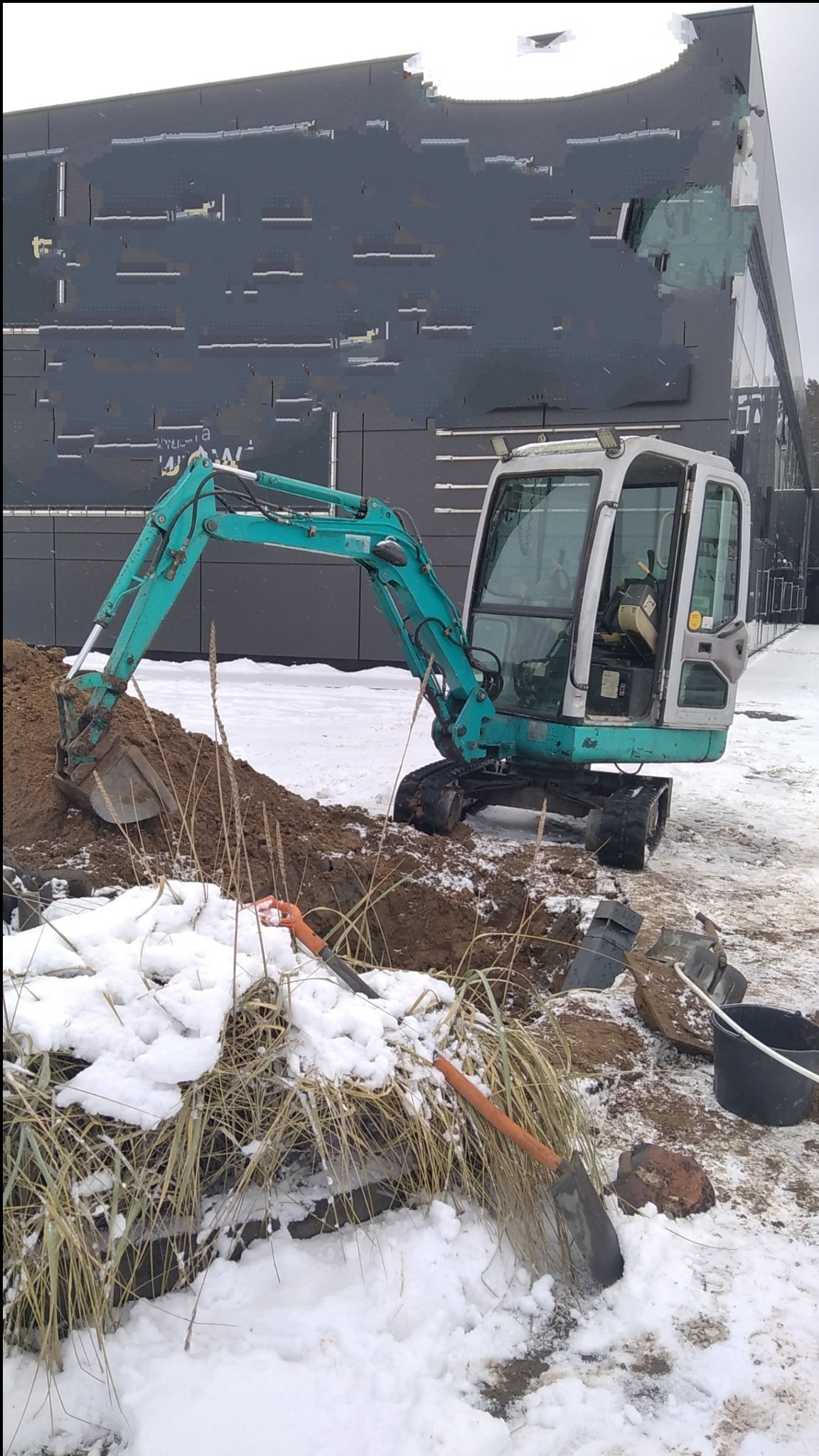 Niebiesko-szara mini koparka na zaśnieżonym placu budowy, obok wykopu, z łopatami i wiadrem, na tle nowoczesnego budynku.