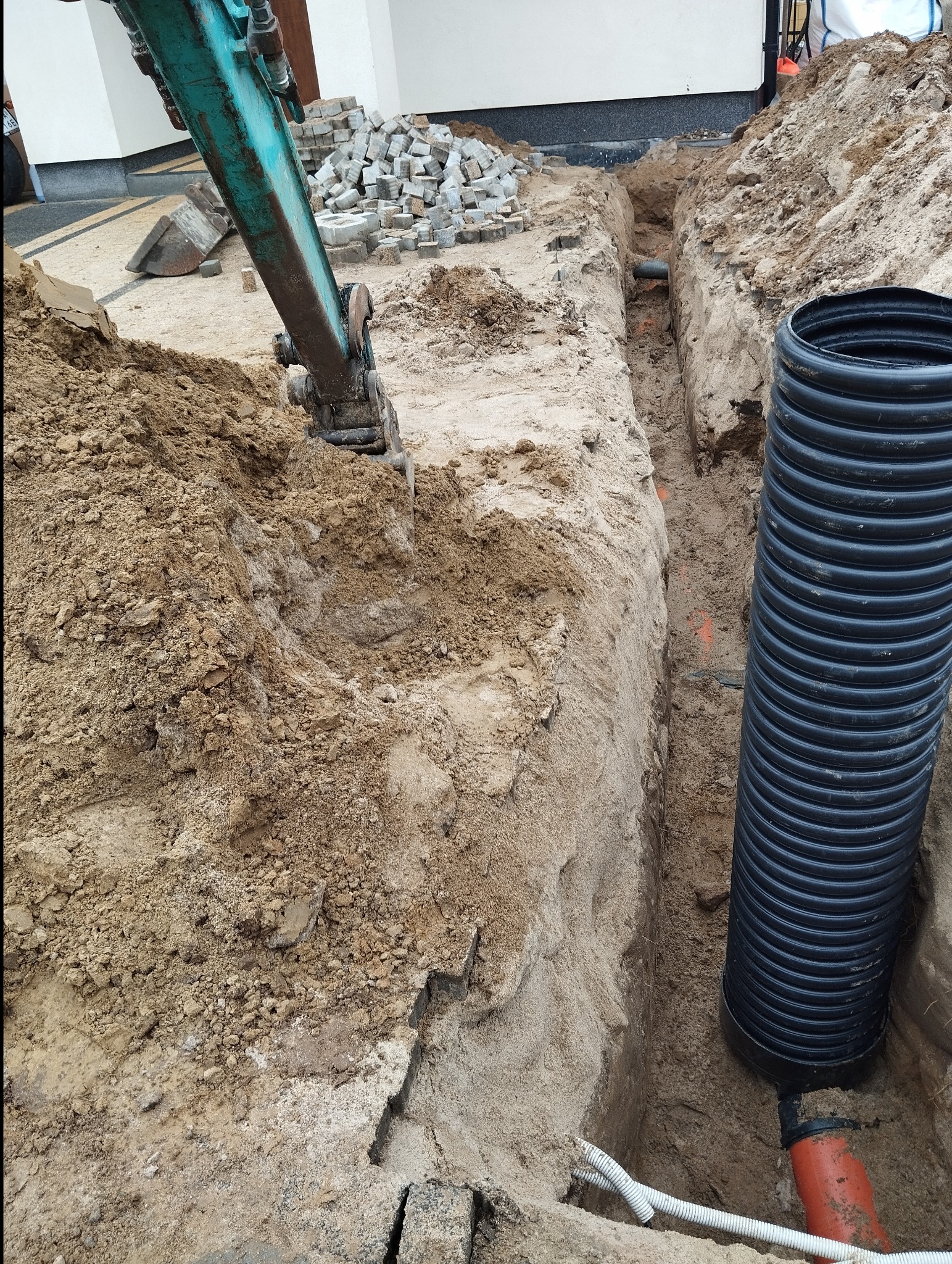 Wykopany wąski rów z ułożonymi rurami kanalizacyjnymi, widoczny fragment koparki i sterta kostki brukowej w tle.