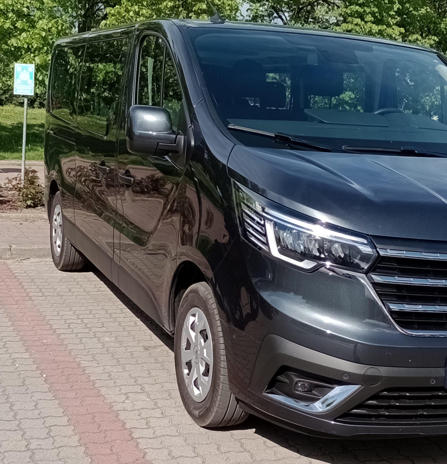 Ciemnoszary, lśniący bus pasażerski zaparkowany na brukowanym placu, z widocznym znakiem drogowym w tle.