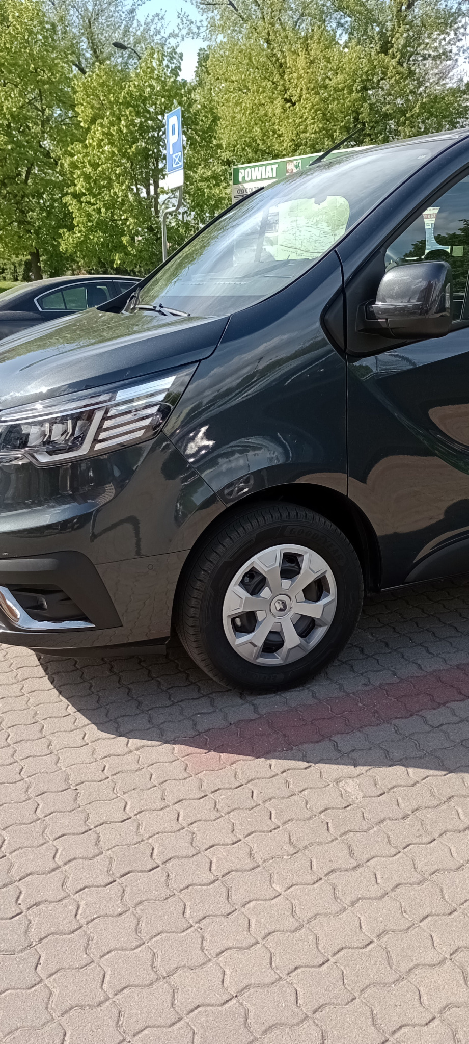 Fragment szarego samochodu typu van na parkingu z kostki brukowej, widoczny fragment koła z srebrną felgą i logo, reflektor oraz tablica informacyjna w tle.