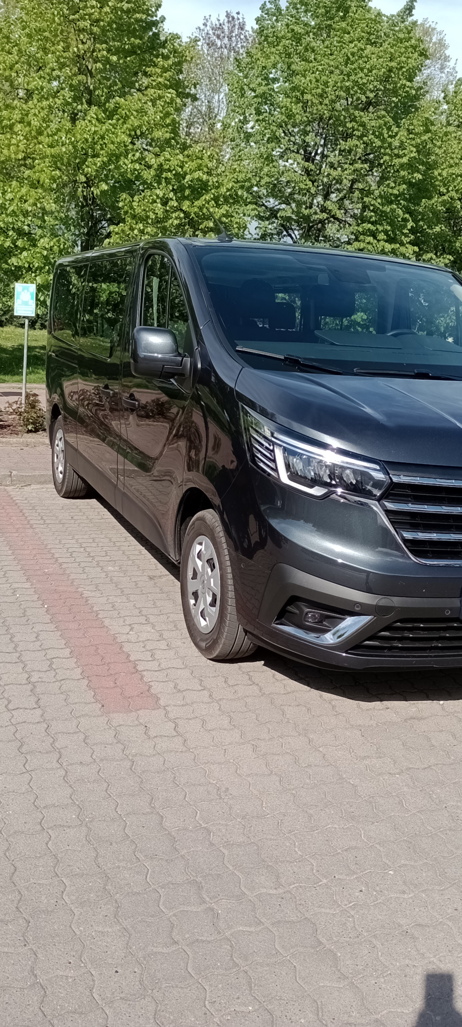 Ciemnoszary van na kostce brukowej z zielonymi drzewami w tle, widok z przodu i boku, delikatne odbicie w karoserii.