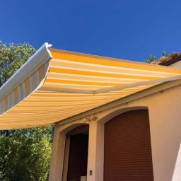 SUNFALL SPÓŁKA Z OGRANICZONĄ ODPOWIEDZIALNOŚCIĄ - Żółto-biała markiza tarasowa z poziomymi pasami, zamontowana nad dwoma oknami z brązowymi roletami zewnętrznymi, na tle błękitnego nieba i zieleni.