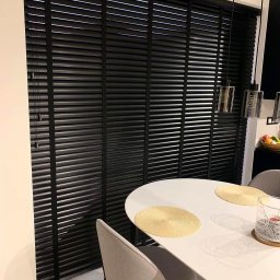 SUNFALL SPÓŁKA Z OGRANICZONĄ ODPOWIEDZIALNOŚCIĄ - Eleganckie, czarne żaluzje wewnętrzne drewniane, zamontowane w oknie nad białym stołem z okrągłymi podkładkami, oświetlone lampami wiszącymi.