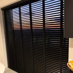 SUNFALL SPÓŁKA Z OGRANICZONĄ ODPOWIEDZIALNOŚCIĄ - Czarne drewniane żaluzje poziome w oknie z widokiem na zachodzące słońce, widoczne z wnętrza pomieszczenia.