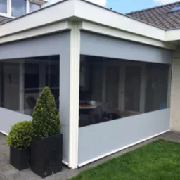 Zadaszony taras z szarymi roletami przeciwsłonecznymi, widok z zewnątrz, z dekoracyjnymi krzewami w donicach.