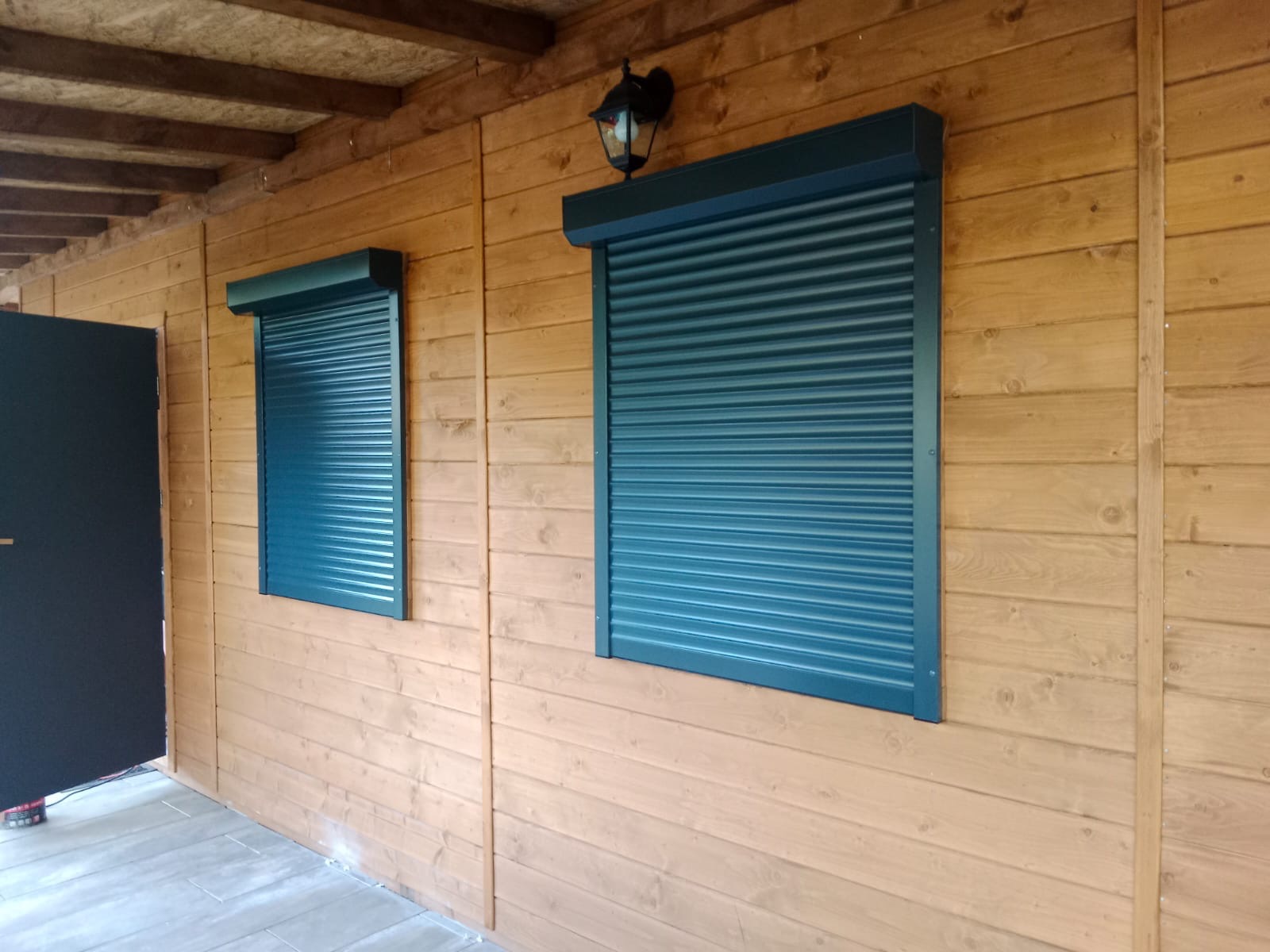 Dwa okna z zamkniętymi, turkusowymi osłonami na drewnianej elewacji domku letniskowego z widoczną lampą zewnętrzną i fragmentem czarnych drzwi.