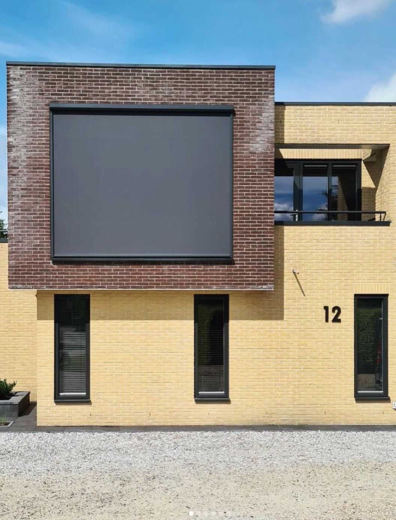 Elewacja domu z żółtej cegły z szarą, dużą osłoną przeciwsłoneczną na ceglanej wstawce, widoczne okna z żaluzjami wewnętrznymi i numer domu 12.
