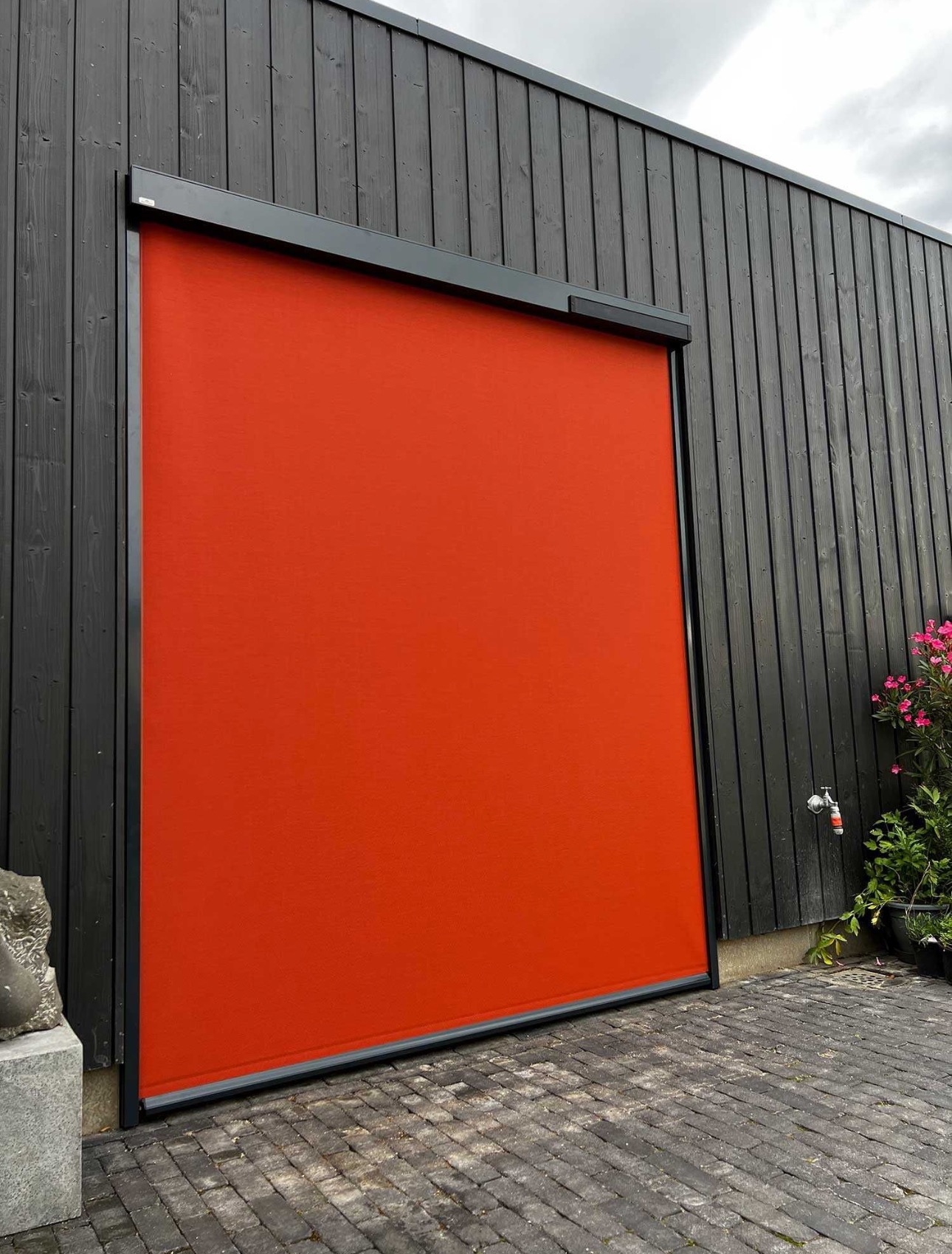 Pomarańczowa osłona przeciwsłoneczna na elewacji budynku z ciemnego drewna, widoczny fragment kamiennej rzeźby i kran z wodą.