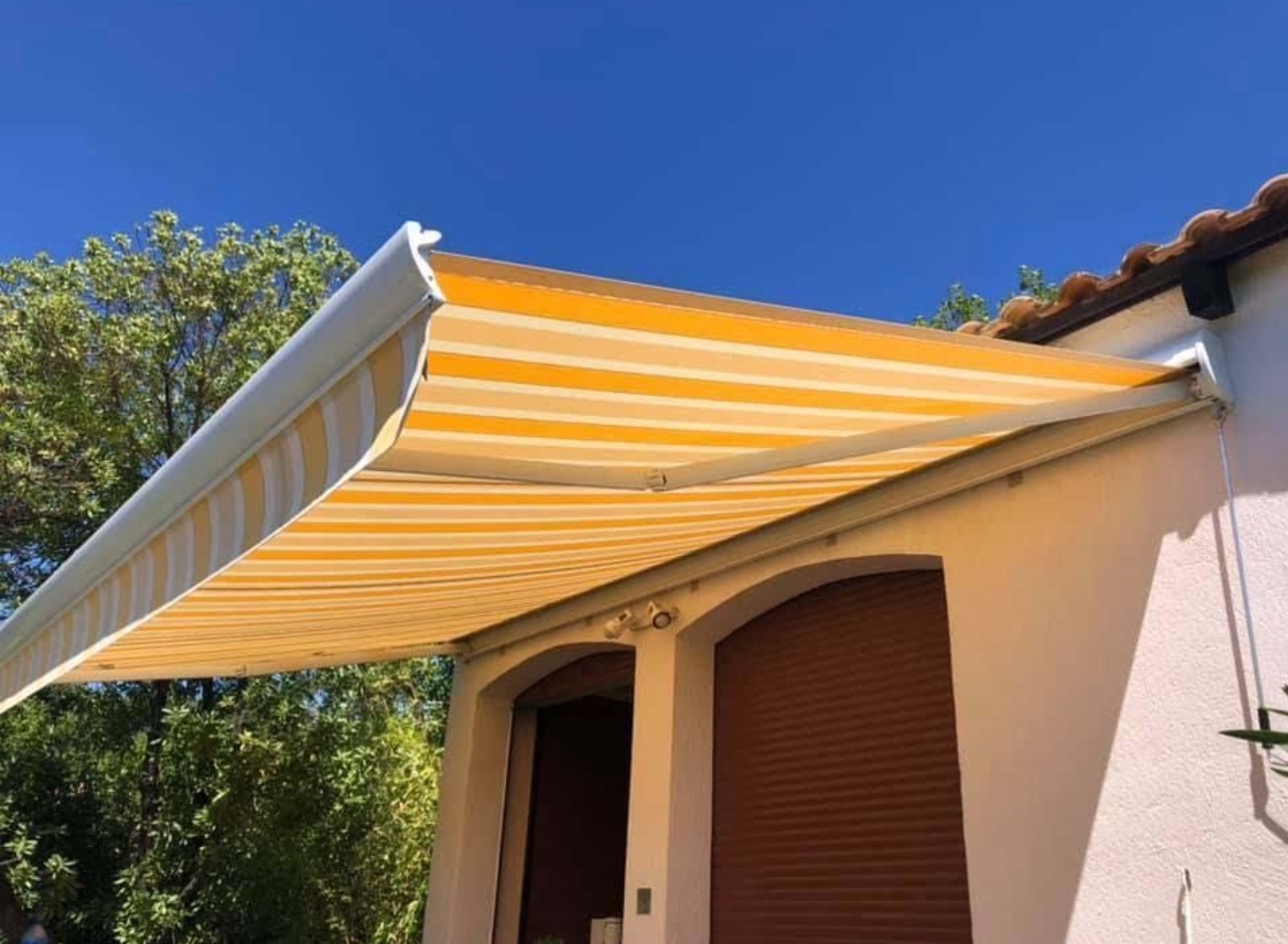 Żółto-biała markiza tarasowa z poziomymi pasami, zamontowana nad dwoma oknami z brązowymi roletami zewnętrznymi, na tle błękitnego nieba i zieleni.