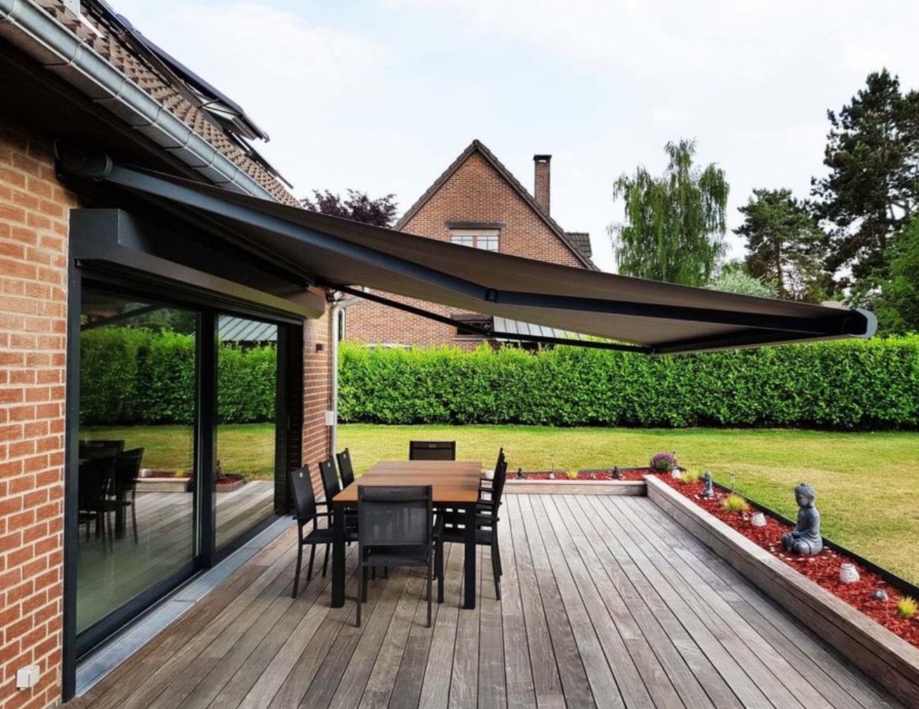 Zewnętrzna markiza tarasowa z szarą tkaniną, zamontowana nad przesuwnymi drzwiami balkonowymi w domu z czerwonej cegły, widok na drewniany taras z meblami ogrodowymi i starannie utrzymany trawnik.