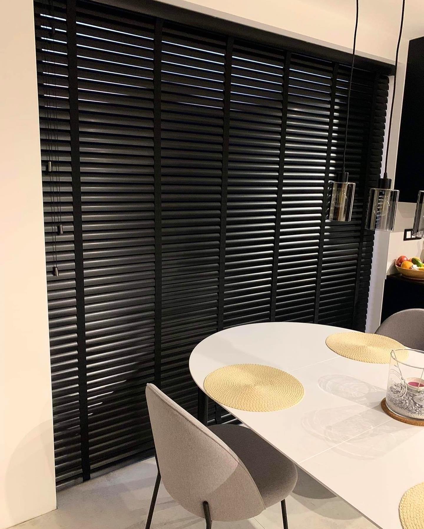 Eleganckie, czarne żaluzje wewnętrzne drewniane, zamontowane w oknie nad białym stołem z okrągłymi podkładkami, oświetlone lampami wiszącymi.