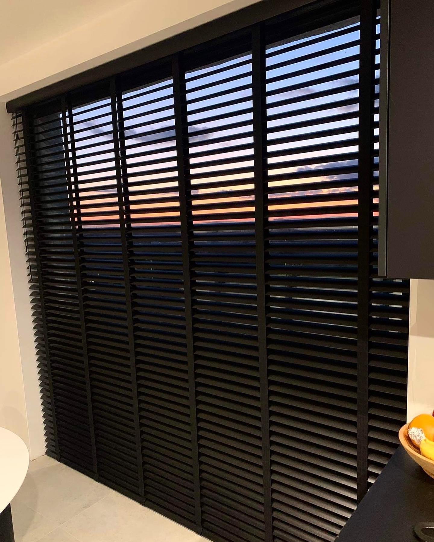 Czarne drewniane żaluzje poziome w oknie z widokiem na zachodzące słońce, widoczne z wnętrza pomieszczenia.