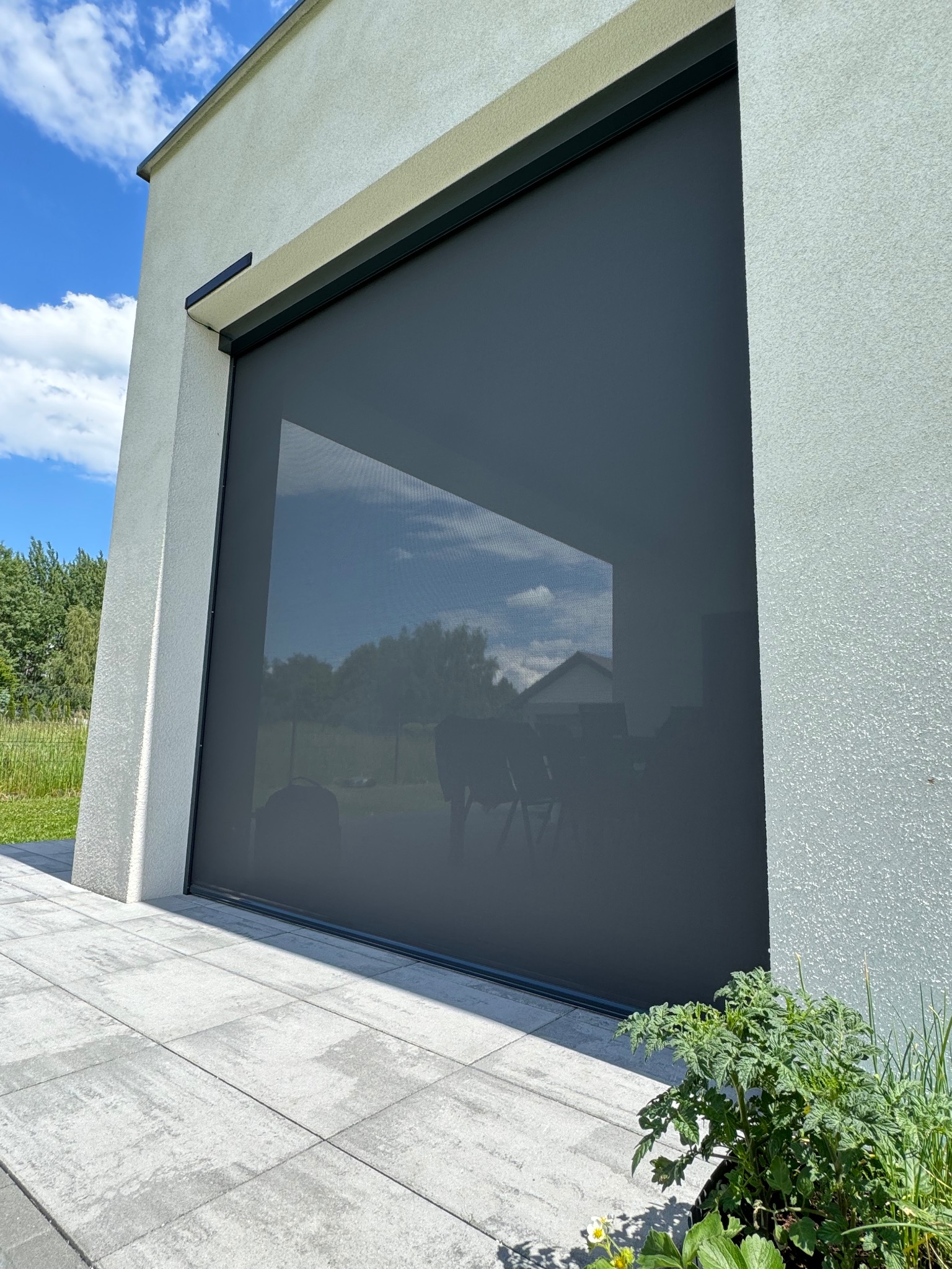 Grafitowa osłona okienna na elewacji budynku, z widocznym odbiciem nieba i domu w strukturze materiału, roślina w donicy u podstawy.