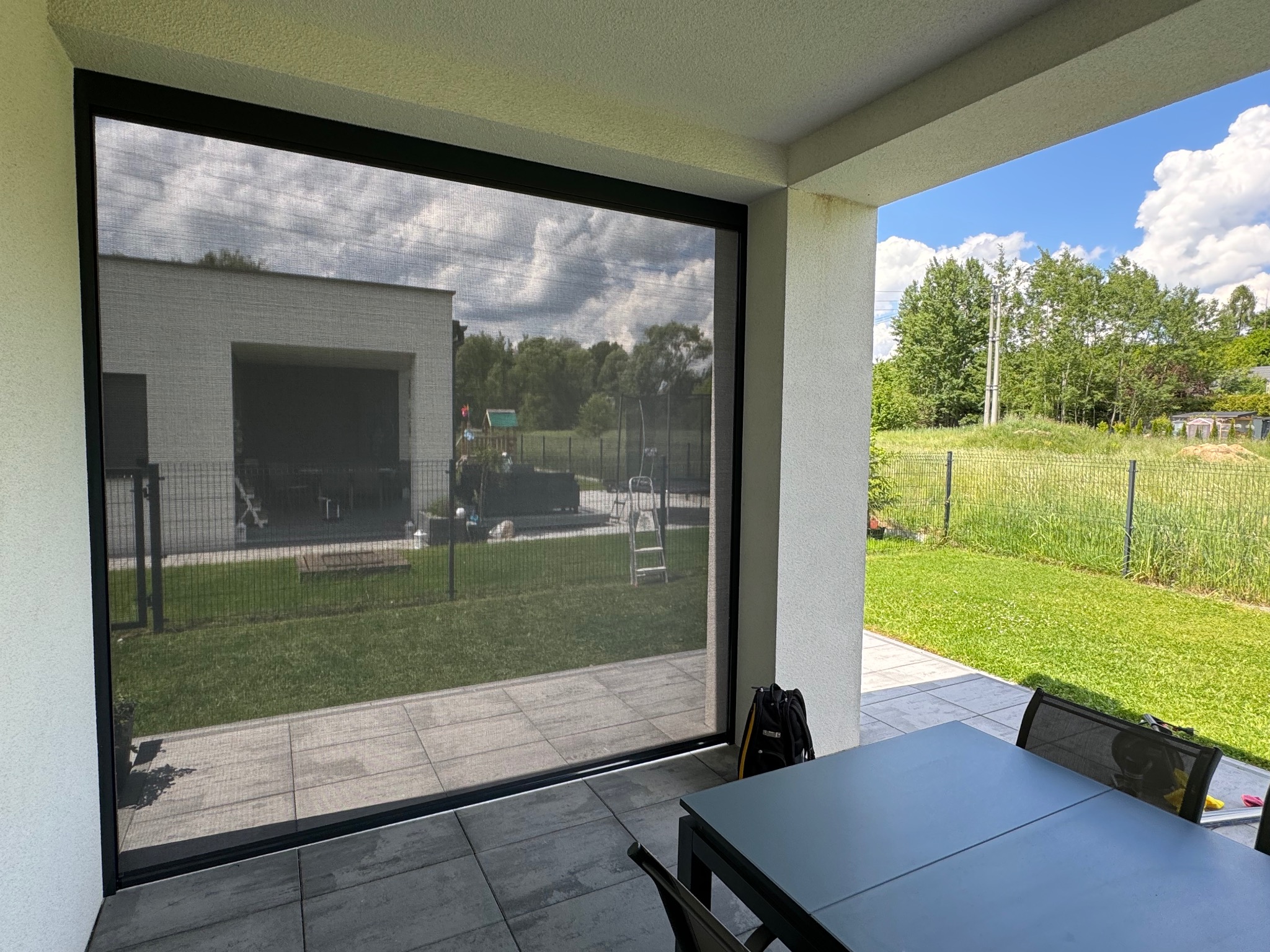 Zewnętrzna moskitiera w czarnej ramie, zamontowana na tarasie, widok na ogród z zielonym trawnikiem i dom w tle.