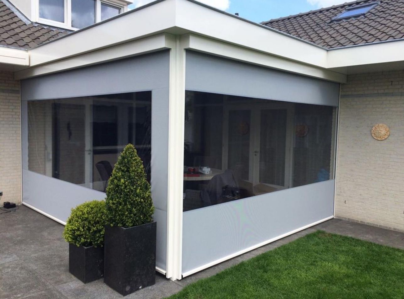 Zadaszony taras z szarymi roletami przeciwsłonecznymi, widok z zewnątrz, z dekoracyjnymi krzewami w donicach.