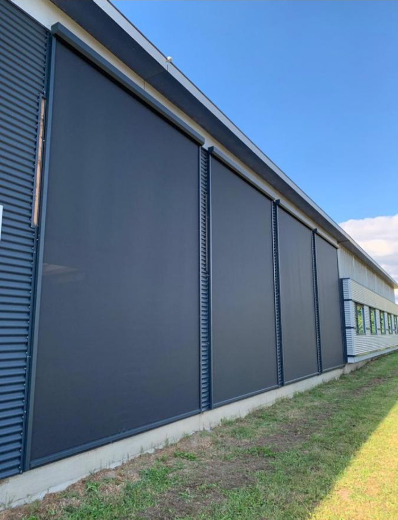 Zewnętrzne, zaciemniające przesłony okienne zamontowane na elewacji budynku przemysłowego z blachy trapezowej, widok z poziomu gruntu.