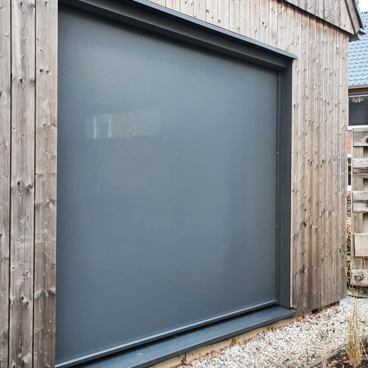 Szara, zwinięta przesłona okienna zamontowana na drewnianej elewacji budynku, widoczne detale konstrukcyjne i otoczenie z kamykami.