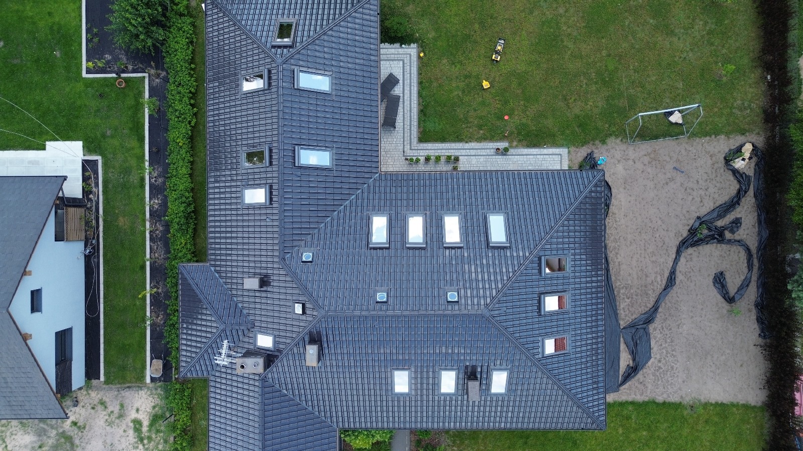 Widok z góry na ciemny dach z licznymi oknami dachowymi, otoczony zielenią i elementami zagospodarowania terenu. Ujęcie z drona.