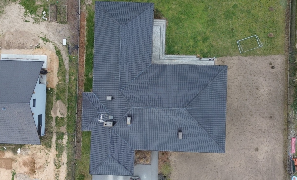 Widok z drona na ciemny, symetryczny dach domu z kominami, otoczony zielenią i piaskiem. Widoczne detale pokrycia dachowego i elementy otoczenia.