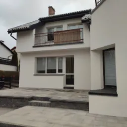 Świeżo otynkowany dom murowany z balkonem i brukowanym tarasem, widok z poziomu gruntu na placu budowy.