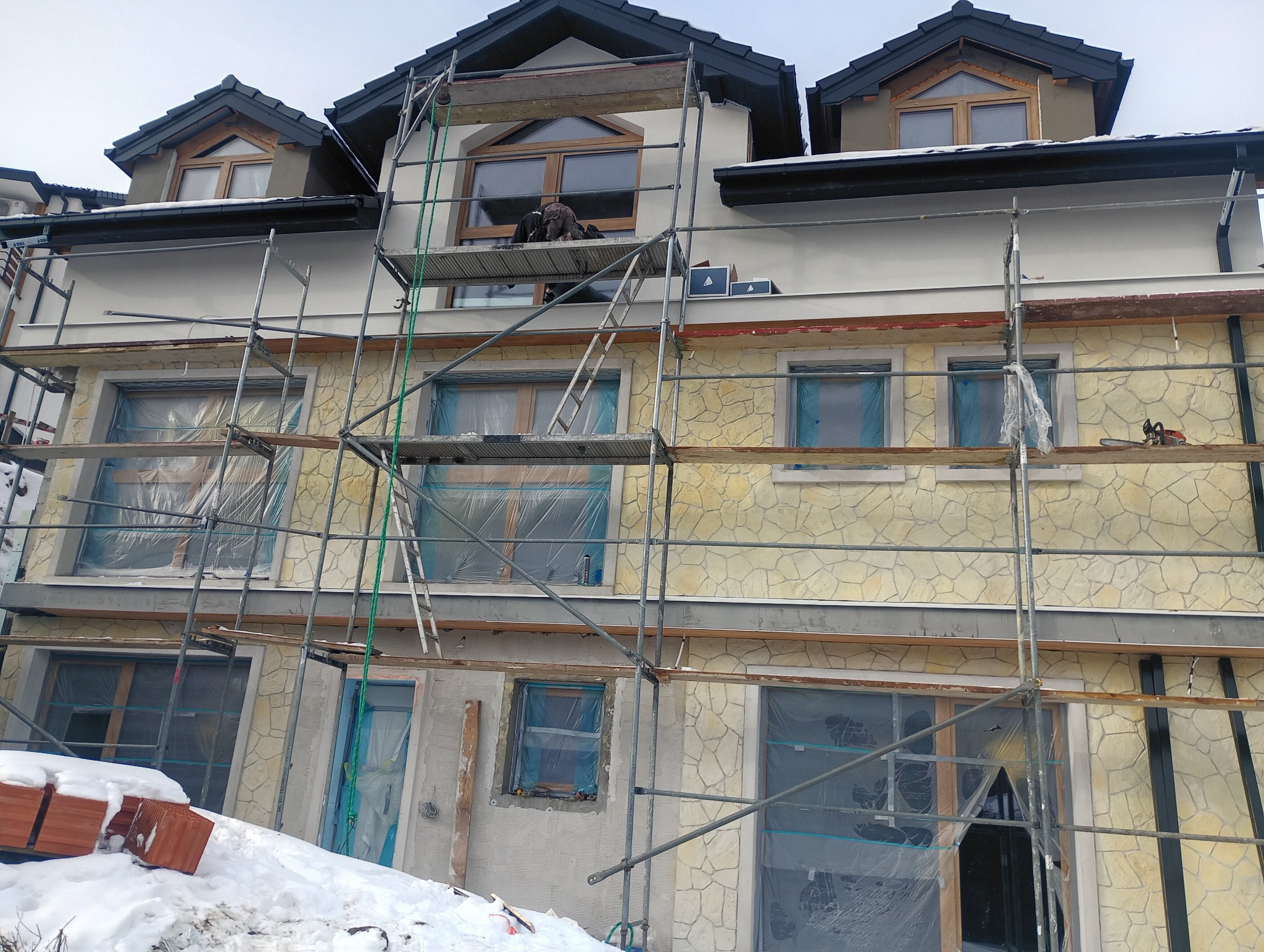 Dwupiętrowy dom w trakcie prac elewacyjnych, z rusztowaniem i zabezpieczonymi oknami. Część elewacji wykończona kamieniem, a część w trakcie tynkowania. Na rusztowaniu pracownik.