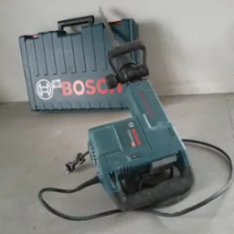 Młot wyburzeniowy marki Bosch z walizką na tle szarej ściany i podłogi.