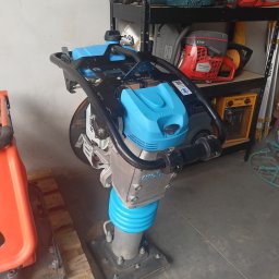 Wypożyczalnia Sprzętu Budowlanego WS-BUD - Nieużywana ubijarka stopowa Wacker Neuson z niebieskimi elementami, stojąca obok pomarańczowej maszyny na drewnianej palecie, z półkami w tle, na których znajdują się piły spalinowe i młot udarowy.