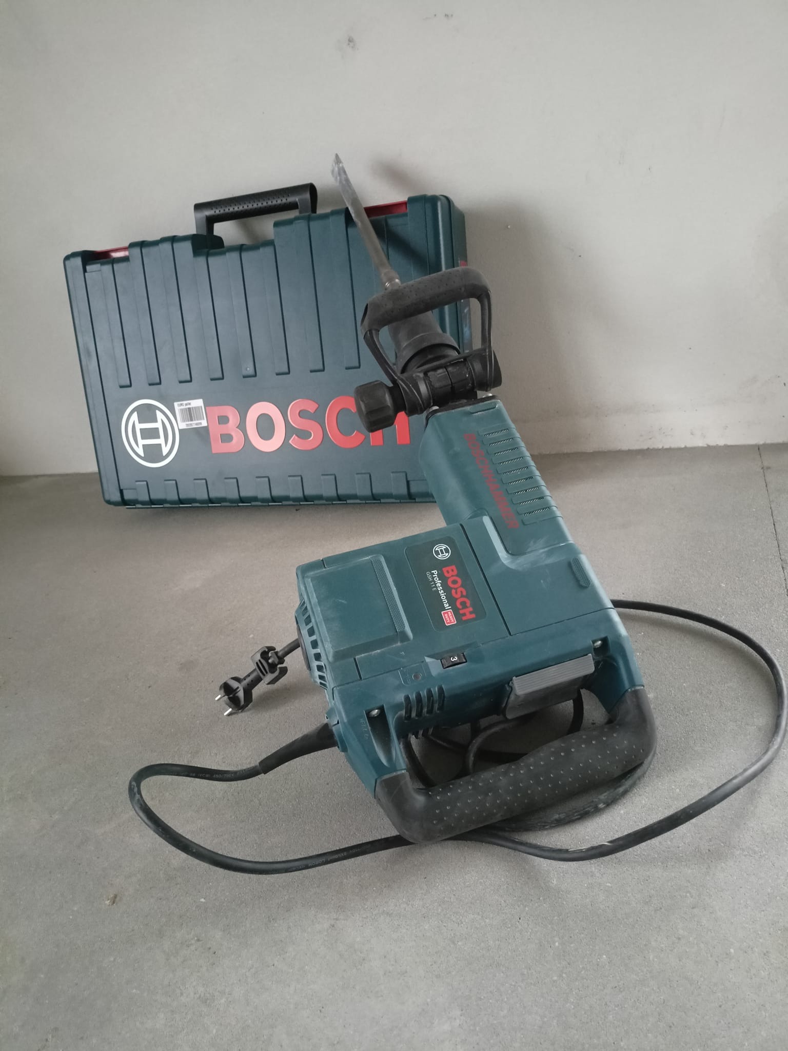 Młot wyburzeniowy marki Bosch z walizką na tle szarej ściany i podłogi.