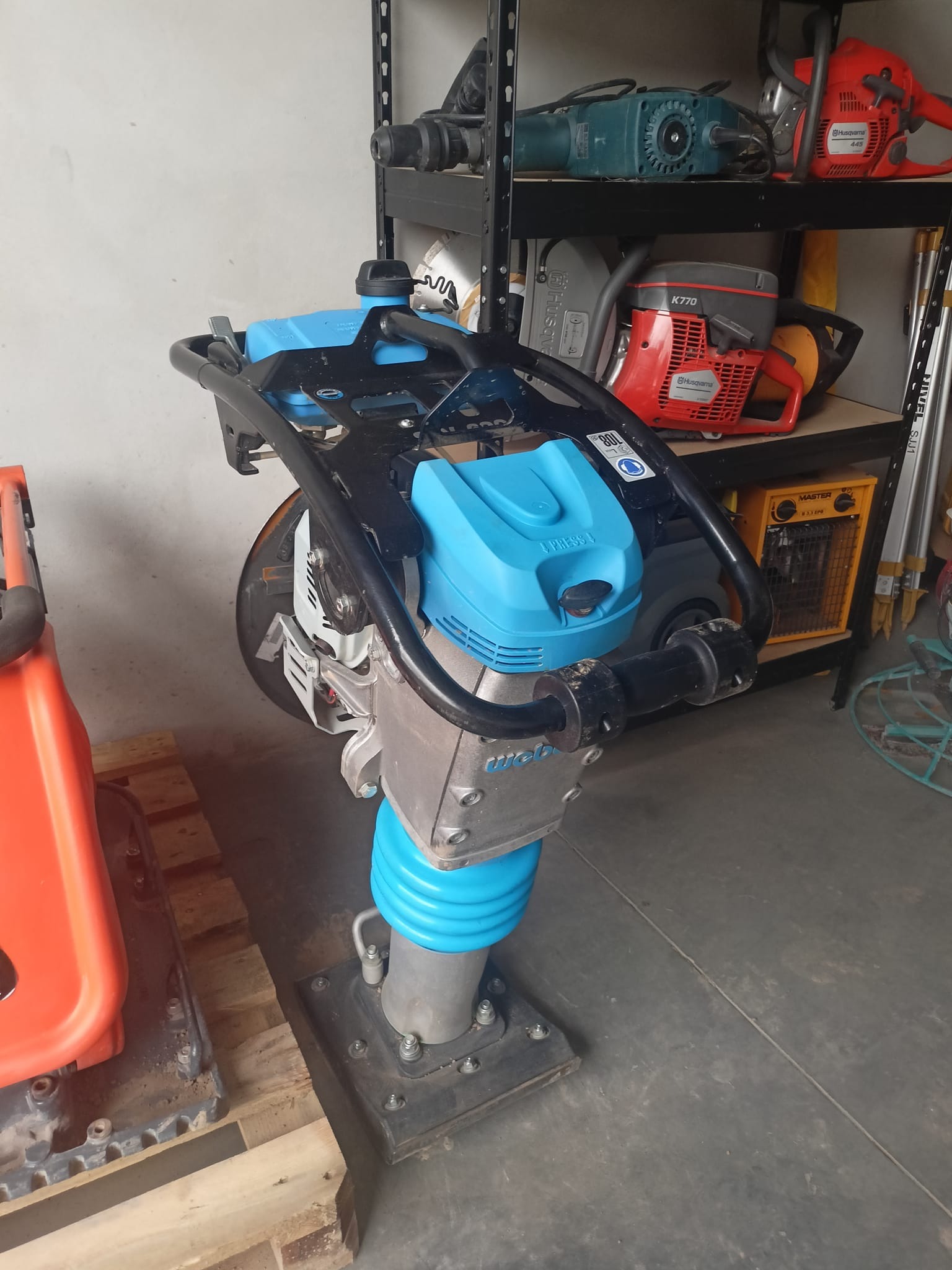 Nieużywana ubijarka stopowa Wacker Neuson z niebieskimi elementami, stojąca obok pomarańczowej maszyny na drewnianej palecie, z półkami w tle, na których znajdują się piły spalinowe i młot udarowy.