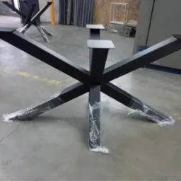 Surowa, czarna, metalowa podstawa stołu o geometrycznym kształcie X, z elementami zabezpieczonymi folią, na szarym, betonowym podłożu w warsztacie.