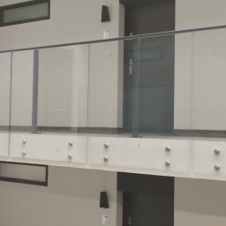 MD Nowoczesne konstrukcje - Szklana balustrada z metalowymi elementami mocującymi, widoczna z perspektywy dolnej kondygnacji, w nowoczesnym wnętrzu budynku z drzwiami i oknami.