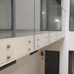 MD Nowoczesne konstrukcje - Szklana balustrada z mocowaniami punktowymi w nowoczesnym wnętrzu. Widoczny rower i drzwi wejściowe w tle. Jasne, neutralne kolory ścian i podłogi.