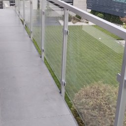 MD Nowoczesne konstrukcje - Nowoczesna balustrada szklana z aluminiowymi słupkami na balkonie z szarymi płytkami. Widok na zadbany trawnik i dom z ciemnym ogrodzeniem.