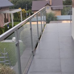 MD Nowoczesne konstrukcje - Nowoczesny balkon z szarą, kwadratową podłogą i szklaną balustradą z aluminiowymi profilami. W tle domy i zieleń. Widok z góry, spokojne oświetlenie.
