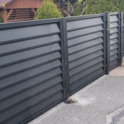 MD Nowoczesne konstrukcje - Nowoczesna, grafitowa brama wjazdowa z poziomymi lamelami, uzupełniona o klasyczne, metalowe ogrodzenie z ozdobnymi elementami. Widoczne detale montażowe.